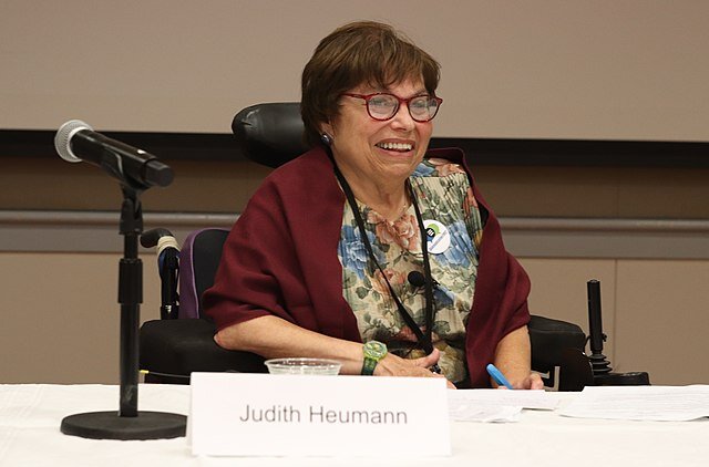 A photograph of Judith Heumann. She is sitting in her power chair behind table covered with a white table cloth. There is a microphone and a name placard in the foreground.